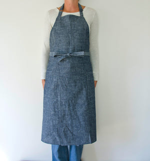 person modeling a split leg apron in linen. they are wearing a white shirt and jeans and standing in front of a white wall. the apron is denim.